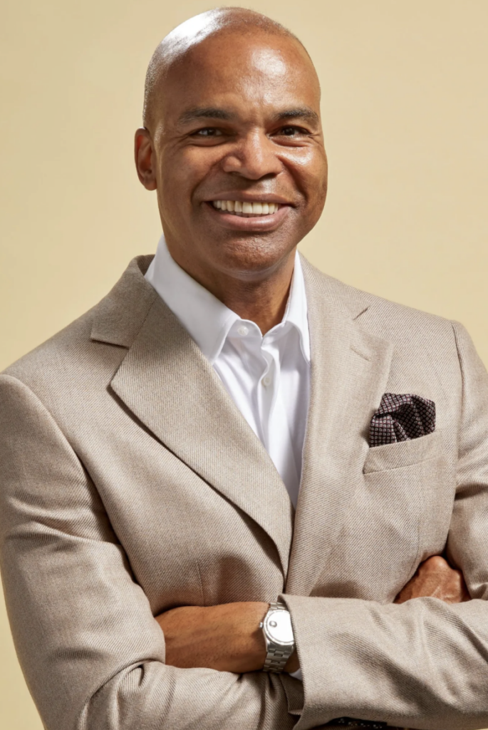 Tommy Amaker wears a suit and smiles against a blank tan background.